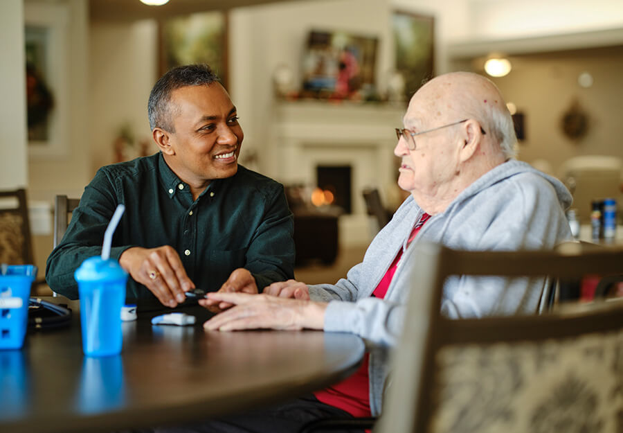 Caregiver assisting a senior man at a round table in a senior living community common area.