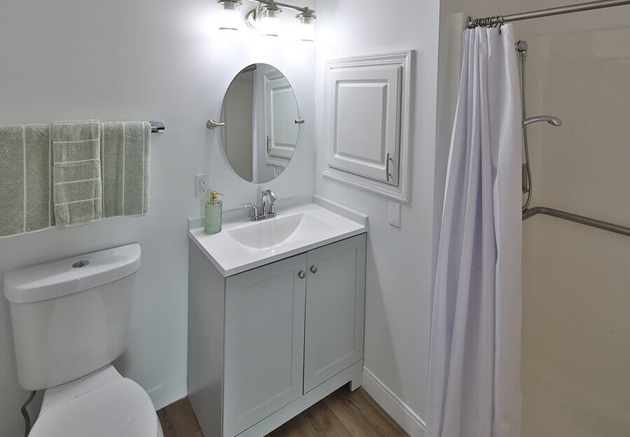 Modern bathroom with white fixtures, round mirror, and shower area in a senior living unit.