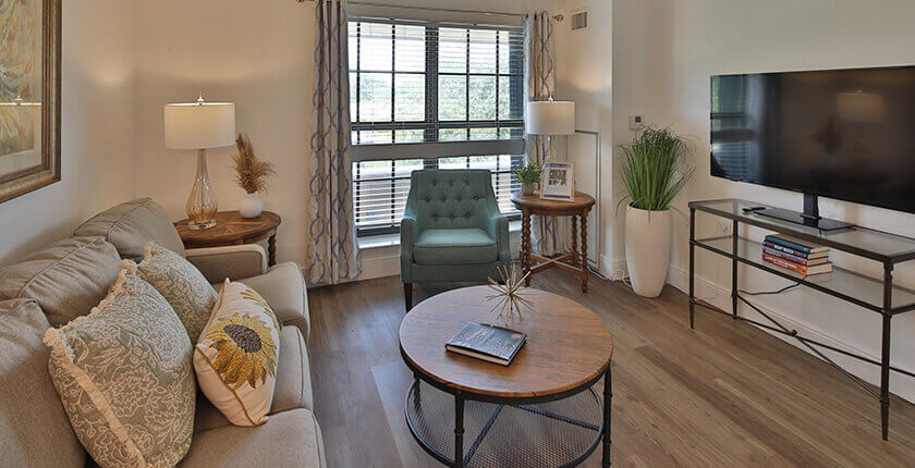 A cozy unit living room with a sofa, coffee table, armchair, and TV in a senior living community.