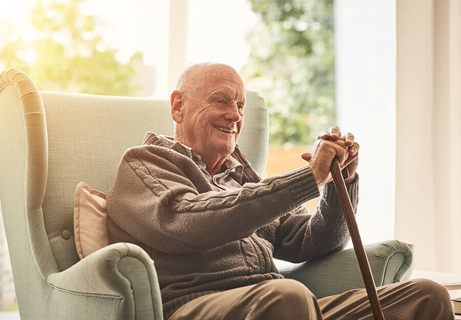 Elderly man smiling while seated in a comfortable armchair, holding a walking stick.