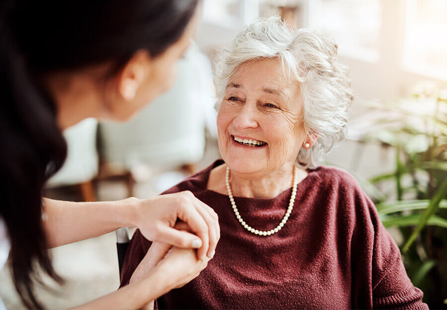 Elderly woman smiling and holding hands with a caregiver in a senior living unit.