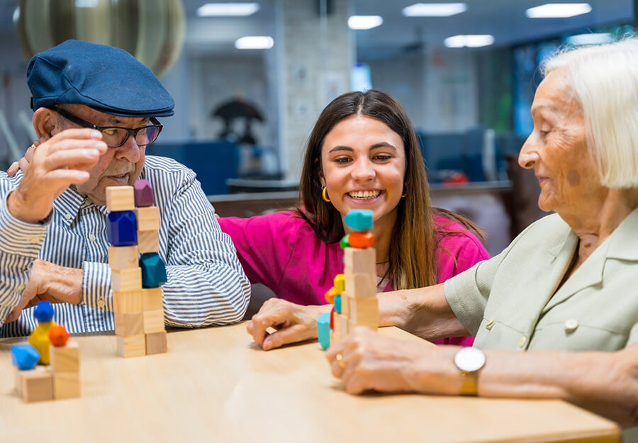 Two senior residents and a caregiver enjoying a game with colored blocks in a community unit.