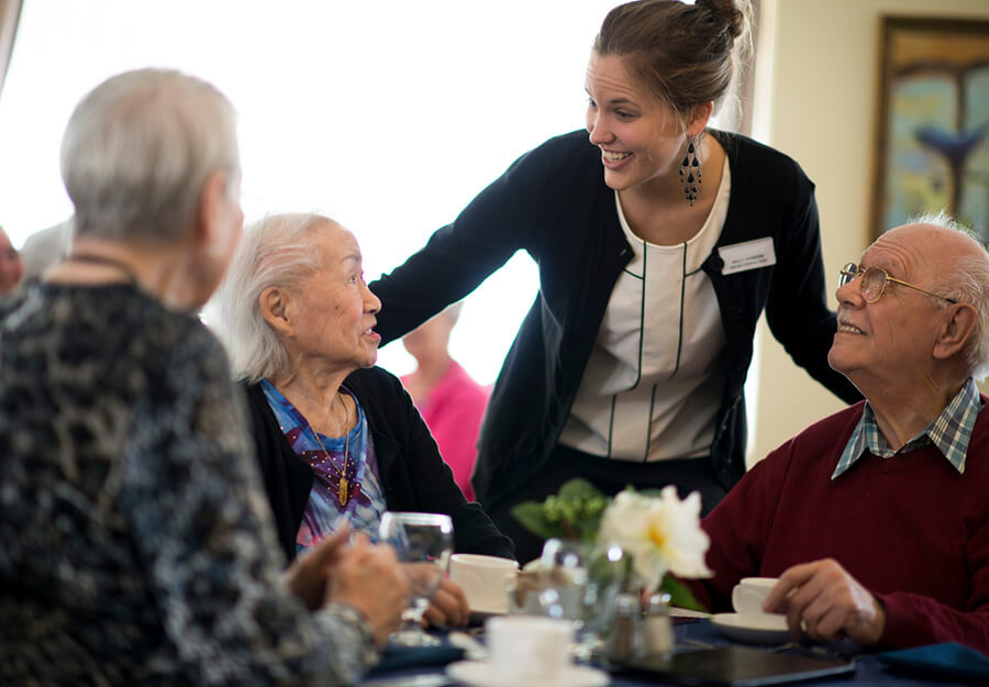 Friendly caregiver interacting with three elderly residents over tea in a senior living community.