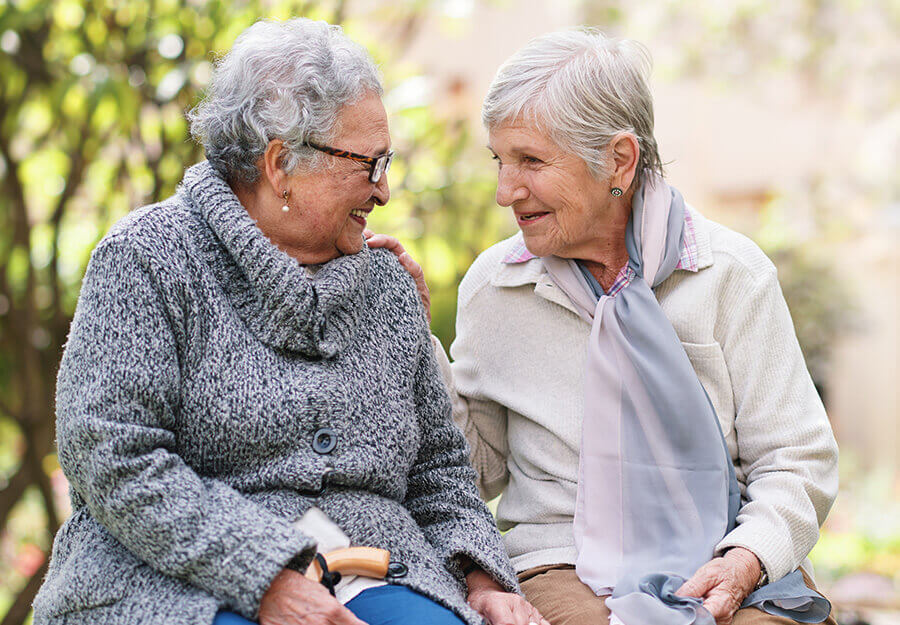 Two elderly women sitting outdoors, smiling and talking to each other on a bench.