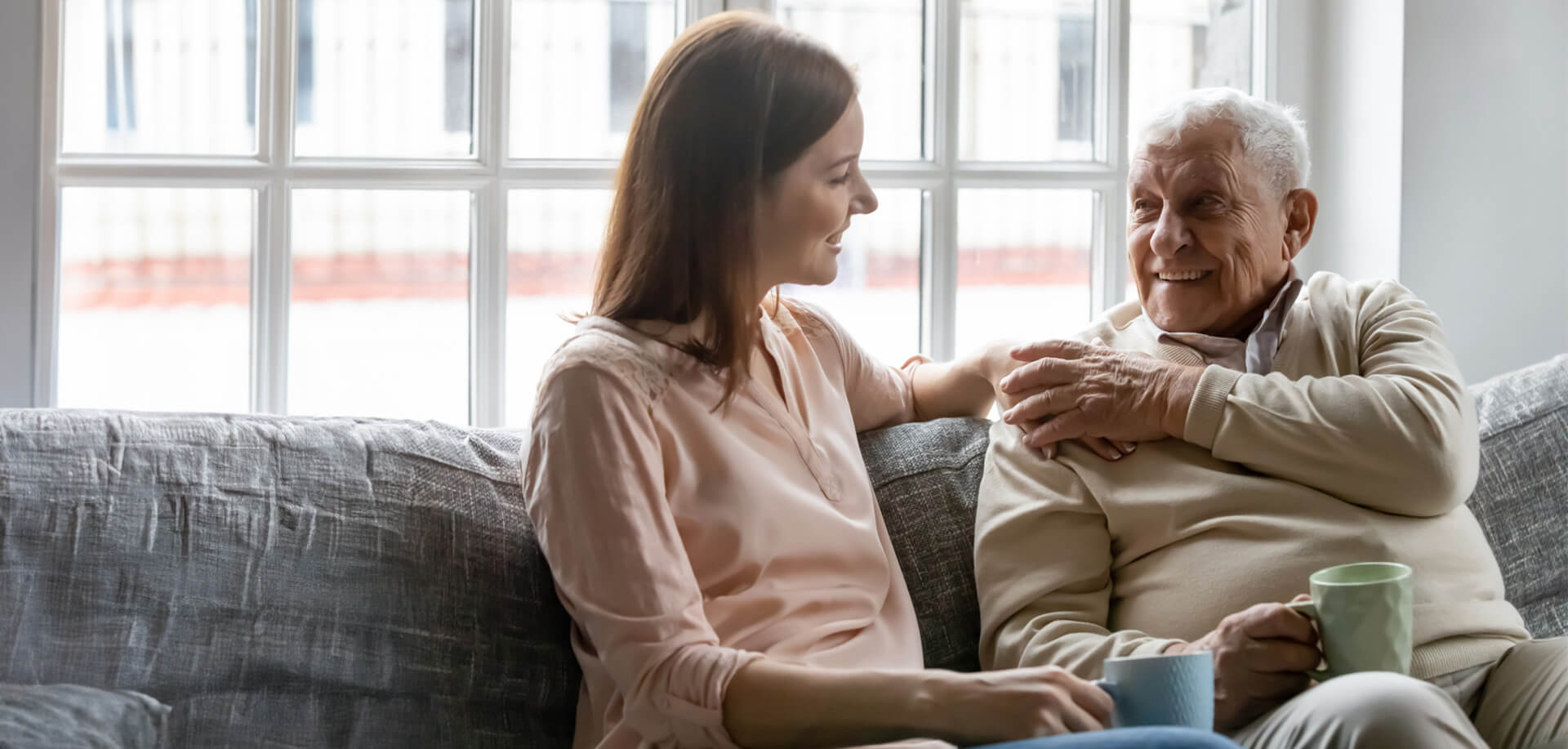 Elderly man and younger woman smiling at each other while sitting on a sofa with mugs in hand.