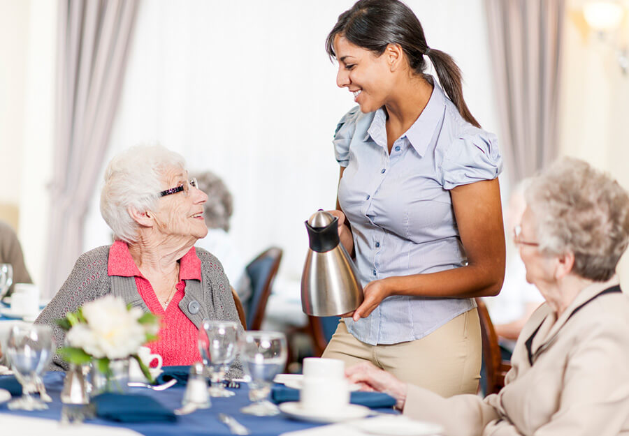 Staff member serves coffee to two elderly women in a senior living dining area.