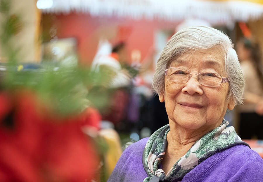 Smiling elderly woman with glasses and purple sweater in a festive living community setting.