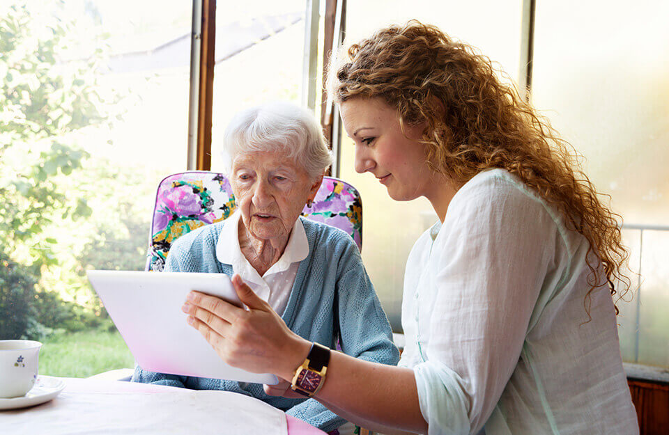 Young woman helping elderly lady navigate a tablet in a bright, cozy unit.