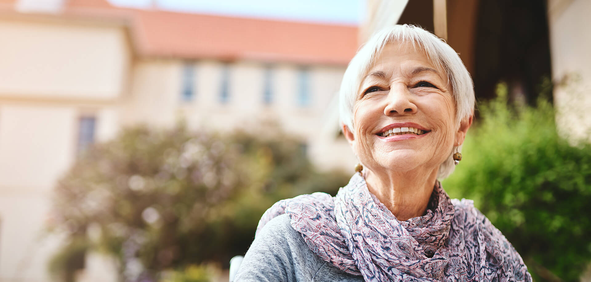 Elderly woman smiling outside a building with a red tile roof and greenery.