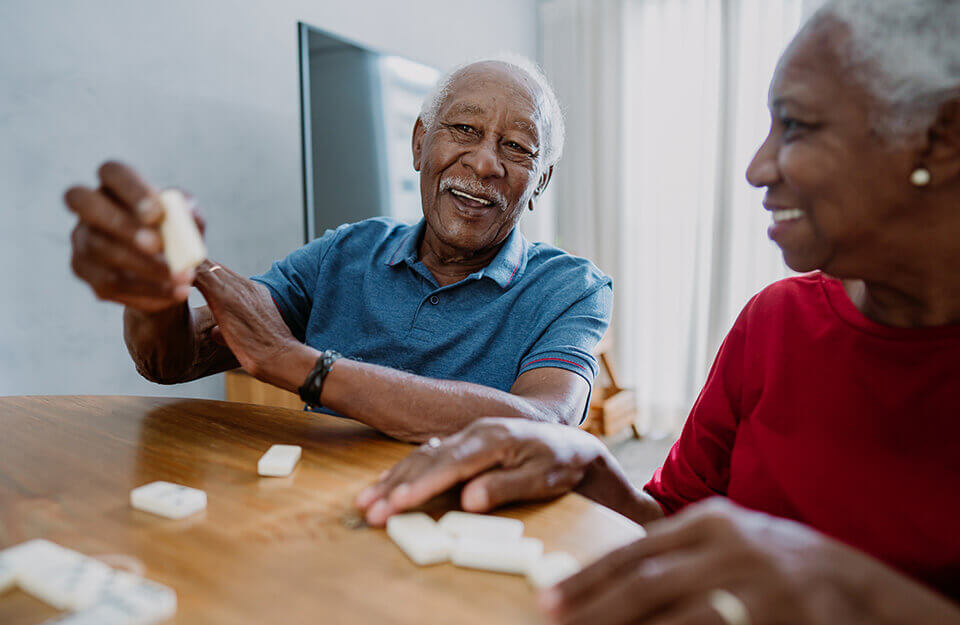 Two seniors happily playing a game of dominoes at a wooden table inside a cozy unit.