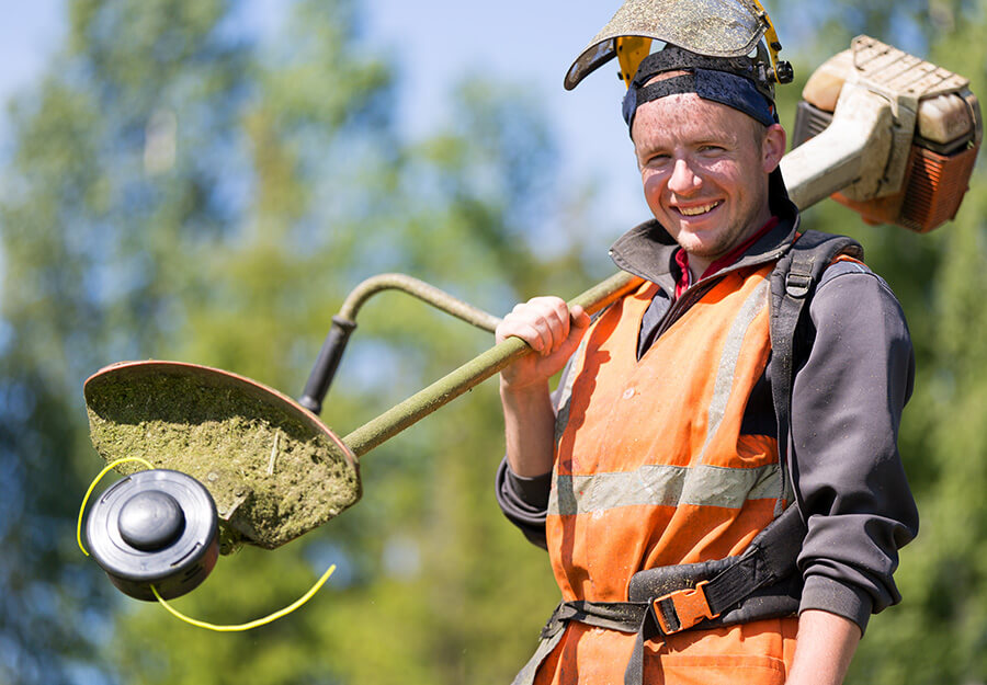 Smiling landscaper holding a weed trimmer, wearing an orange safety vest and helmet outdoors.