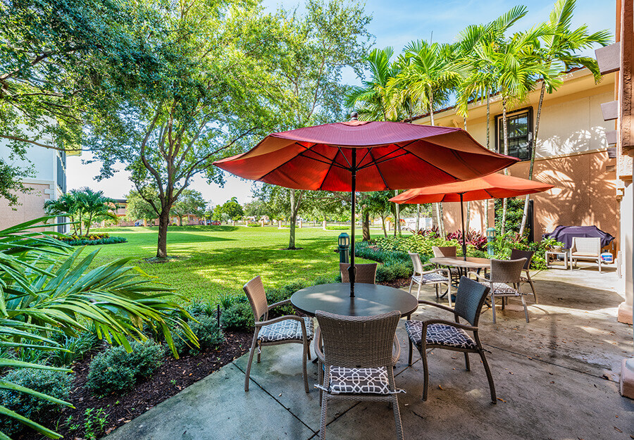 Outdoor area with tables, red umbrellas, and chairs at a senior living community and a lawn.