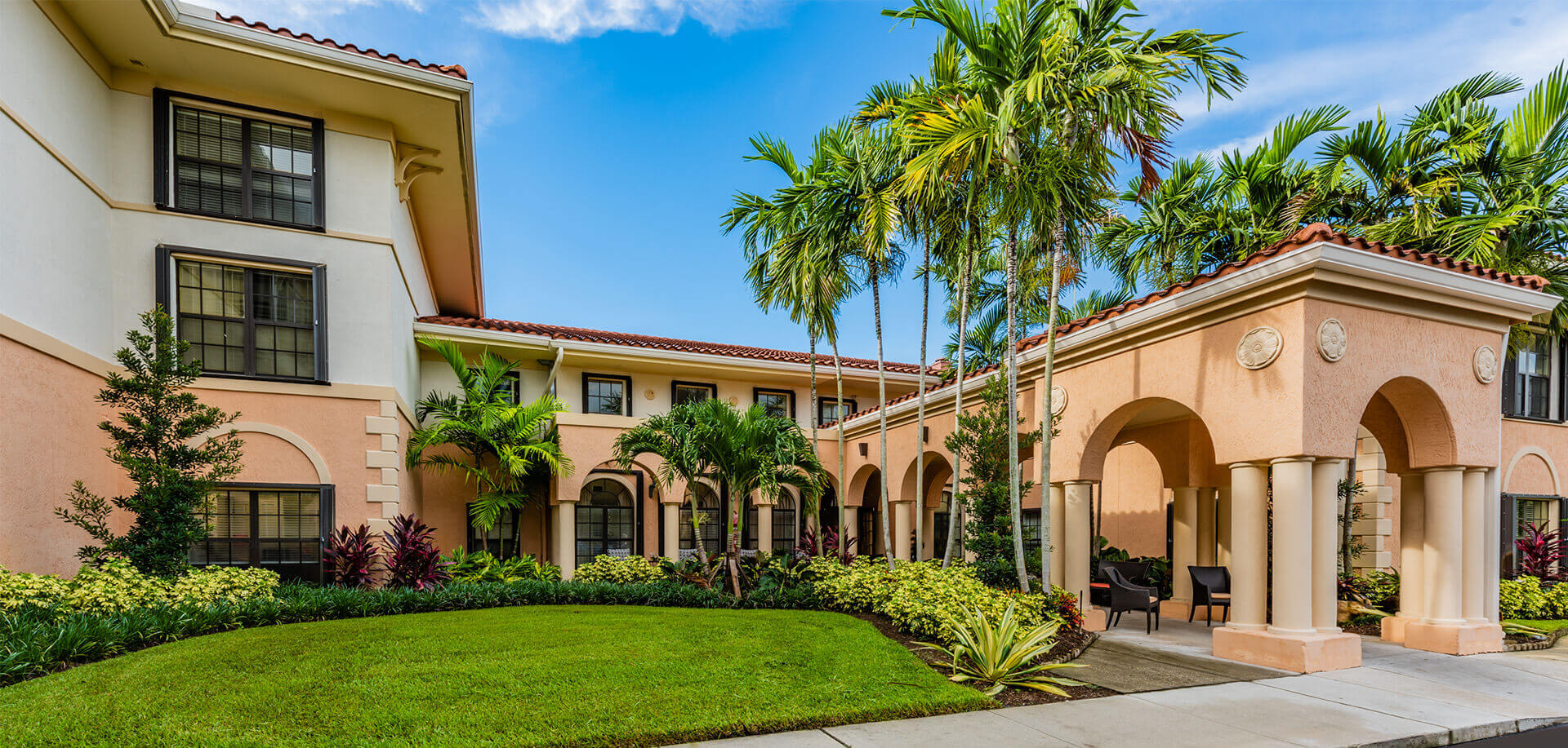 Exterior view of a senior living community building with palm trees and well-manicured lawn.