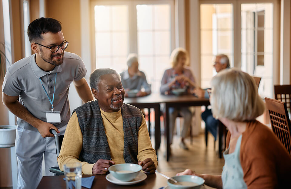 Caregiver assisting elderly residents during a meal in a communal dining area of a senior living community.