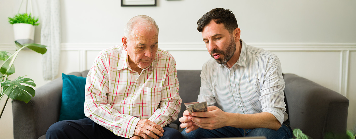 Senior man and younger man sit on a couch together and review finances
